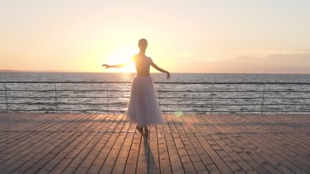 Gyönyörű fiatal balerina, fehér szoknyában sziluettje. Ennek klasszikus balett mozog. Töltés közelében a tenger, vagy óceán. Süt a nap, a tenger felett — Stock videók