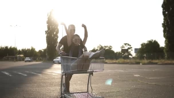 Happy young couple having fun riding outdoors on a parking lot. Man pushing shop cart with a girl inside, she is lauging, exciting and raising hands up. Sun shines — Stock Video