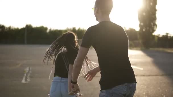 Joven y alegre pareja divirtiéndose juntos en una zona de aparcamiento. Tomados de la mano, girando, riendo. El uso de pantalones cortos de mezclilla y camisetas negras. El sol brilla en el fondo — Vídeo de stock