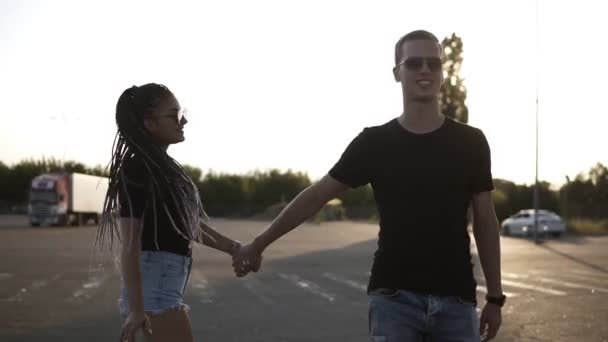 Pareja joven morena rastas chica en gafas de sol y su novio caminando, tomados de la mano. El sol brilla en el fondo — Vídeos de Stock