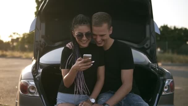 Um casal lindo e jovem a passar tempo juntos. Sentado no porta-malas do carro aberto, abraçando e olhando para o smartphone. Ambos em camisetas pretas, sorrindo, felizes — Vídeo de Stock