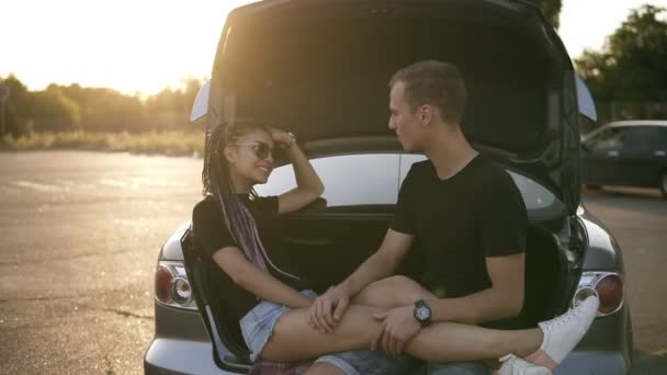 Joven feliz hombre y mujer están sentados juntos en el maletero del coche abierto. Sonrientes y alegres parejas se acarician, riéndose. El sol brilla en el fondo — Vídeo de stock