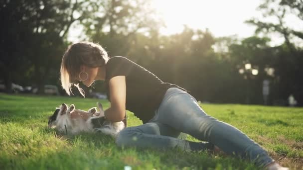 Imágenes frontales de una mujer caucásica en jeans y camiseta negra acostada en la hierba verde del parque y jugando con su mascota. Mujer adulta divertirse con su perrito blanco y negro — Vídeos de Stock