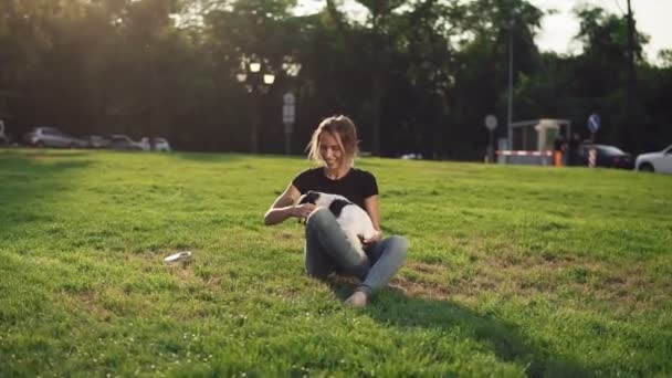 Hermosa mujer joven en casual sentado en la hierba con su mascota perro y sonriendo. Propietario con un perrito encantador al aire libre. Cielo azul y sol en el fondo y hierba verde — Vídeos de Stock