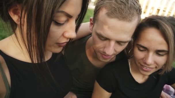 Extremadamente cerca de tres amigos caucásicos: dos mujeres, un hombre joven y un perro pequeño posan para la cámara para tomar fotos juntos. Los amigos están al aire libre en el parque. — Vídeo de stock