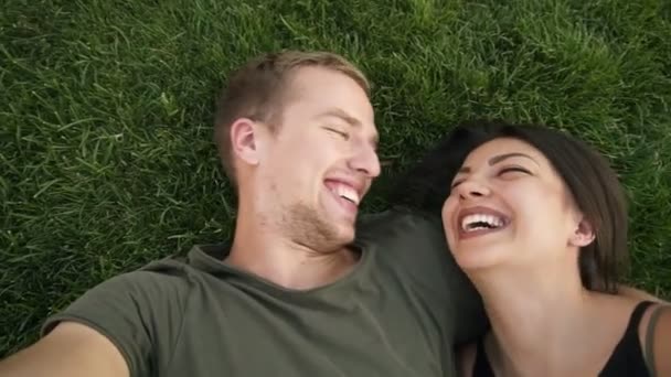 Jóvenes enamorados pareja heterosexual caucásica descansando al aire libre en el parque en el césped, hierba verde acostado en abrazo y haciendo foto selfie en la cámara del teléfono inteligente. Rubia feliz hombre besando a su novia. Parte superior — Vídeos de Stock