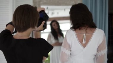 Güzel genç hamile foto model beyaz sabahlığı studio aynaya bakarak kadın fotoğrafçısına poz içinde. Arka görünüm