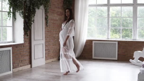 Senhora grávida feliz no vestido branco dançando, girando em torno de sala brilhante. Linda, morena na expectativa. Quarto brilhante com decoração em tijolo — Vídeo de Stock