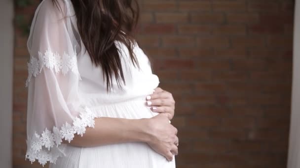 Крупним планом кадри довговолосий брюнетка вагітні моделі позують в білих negilee. Красива жінка кавказька пестити її живіт. Brickwall на тлі — стокове відео