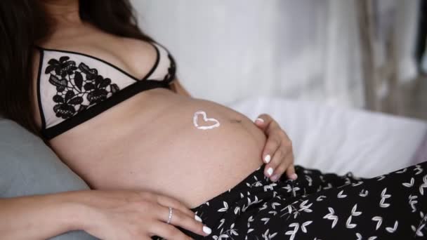 Nahaufnahme eines schönen schwangeren Mädchens in wunderschönem BH, das auf einem Bett liegt, mit Liebessymbol auf dem Bauch der werdenden Mutter. Sie streichelt ihren Bauch mit einer Creme — Stockvideo