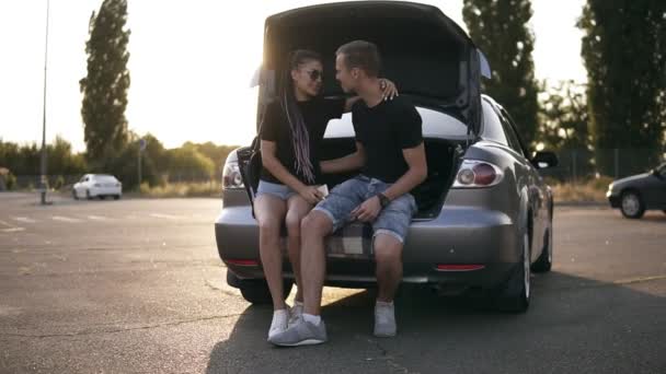 Улыбающаяся пара, сидящая в багажнике современной серебряной машины на парковке. Разговаривали, проводили время вместе. Летнее время — стоковое видео