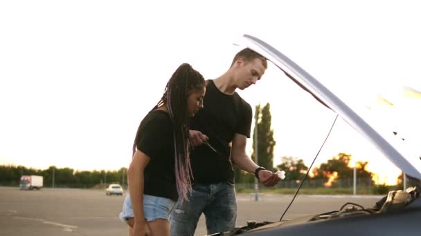 Vista lateral de um jovem casal parou enquanto sua viagem na zona de estacionamento. Jovem de t-shirt preta verifica o nível do óleo, sua namorada com dreadlocks de pé perto do capô do carro aberto. Estacionamento — Vídeo de Stock