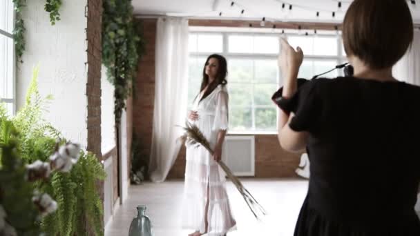 Filmación desde atrás. Fotógrafa en vestido negro tomando fotos de una hermosa mujer embarazada. Habitación luminosa y decorada con ventanas — Vídeos de Stock