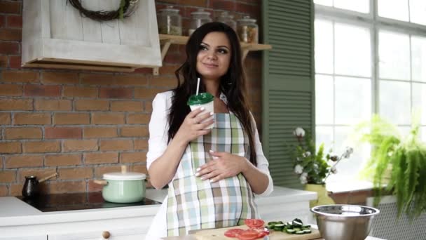 Вид спереди на беременную женщину, трогающую живот на кухне. Белая женщина в кухонном фартуке пьет коктейль из пластиковой чашки с соломинкой. Счастливой семейной жизни — стоковое видео