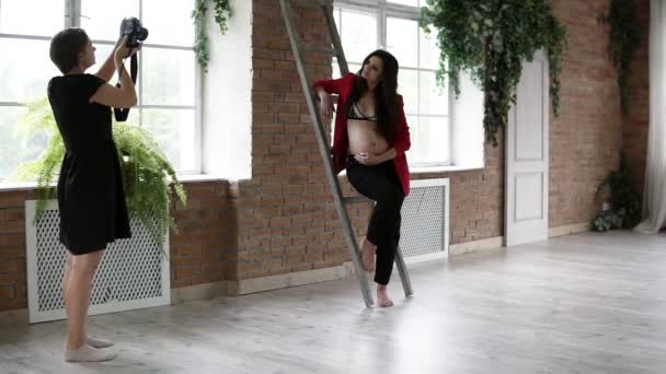 Proceso de fotografía de una hermosa mujer embarazada. Fotógrafa en estudio tomando fotos de una hermosa mujer caucásica con chaqueta roja posando para la cámara apoyada en las escaleras. En interiores — Vídeo de stock