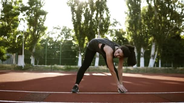 Atleta sexy esbelto executa encostas para baixo em pernas pretas. Exercícios para fortalecer os músculos dos quadris e pernas. Pequeno estádio ao ar livre — Vídeo de Stock