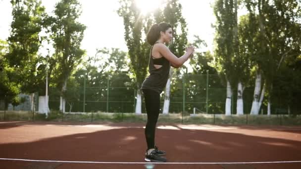 Jonge vrouw atleet in de sport kleding squat oefening buiten. Spectaculaire brunette in zwart T-shirt en legging enthousiast geliefde lichaamsbeweging te doen. Zijaanzicht — Stockvideo