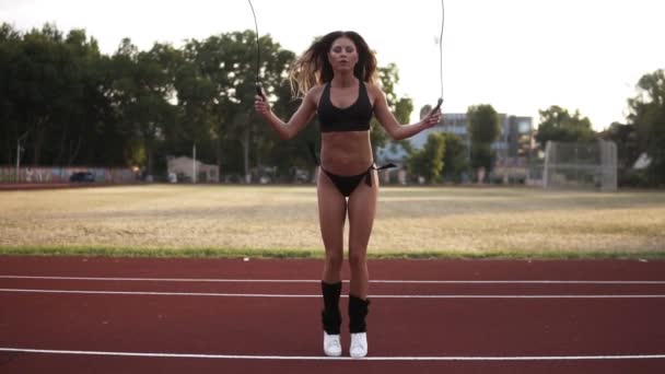 Slow motion van fit, gespierd vrouw in zwarte bikini springen, met behulp van springtouw in openlucht stadion. Vooraanzicht — Stockvideo