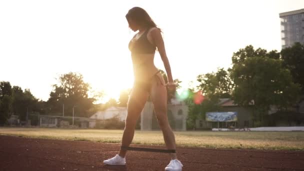 Young sporty model in bikini doing deep squats and sit ups with resistance band outdoors. Sun shines on the background — Stock Video