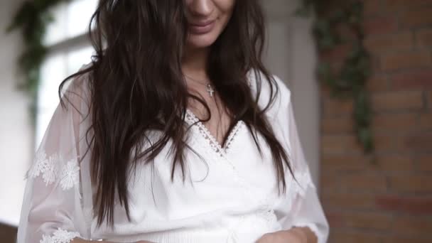 Женщина держит свои руки в форме сердца на своем беременном животе. Беременный живот, женщина в белом платье в помещении — стоковое видео