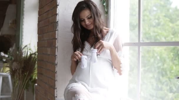 Pregnant, beautiful woman sitting on the sill by the window. Holding babys booties or socks, making little steps on her pregnant belly — Stock Video