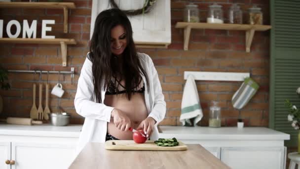 Schöne kaukasische schwangere Frau mit dunklen langen Haaren, die Gemüse auf einem Schneidebrett in der modernen, dekorierten Küche schneidet. Frontansicht — Stockvideo