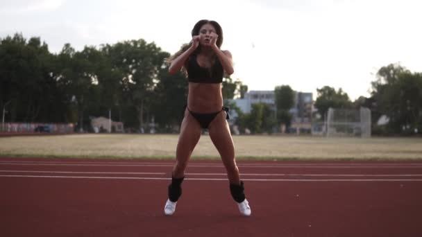 Vältränad ung kvinna gör längd hoppar utomhus på stadion. Närbild av muskulös kvinna i svart bikini tränar. Framifrån — Stockvideo