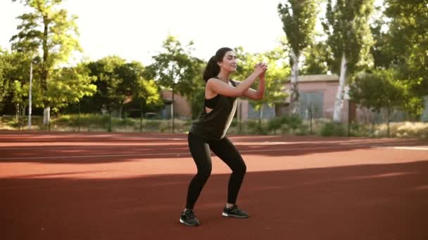 Filmagem em câmera lenta de comprimento total de uma garota morena bastante apta em sportswear fazendo agachamentos enquanto estava em um chão marrom no estádio ao ar livre — Vídeo de Stock