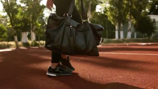 Frau in schwarzen Leggings und mit schwarzer Sporttasche in der Hand läuft an den Gleisen des Stadions entlang. Sporttasche - zielgerichtetes Filmmaterial — Stockvideo