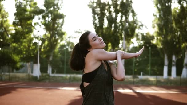 Isınma kadar bir sportif esmer kız onun elleri muscules germe spor giyim önden görünümü. Arka plan açık havada Stadyumu, yeşil ağaçlar — Stok video