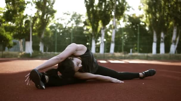 Молодая брюнетка в спортивной одежде сидит на коричневом полу на открытом стадионе, растягивая ноги, делая бечевку. Тренируйся, будь здоров и здоров — стоковое видео