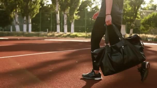 Joven bonita mujer fitness que lleva bolsa de deporte mientras camina por el estadio al aire libre. Sin cara. El sol brilla en el fondo — Vídeos de Stock