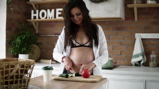 Здоровое питание, беременность, питание и концепция людей - беременная брюнетка в бюстгальтере и белой рубашке, готовящая овощной салат и рубящий огурец на домашней кухне — стоковое видео
