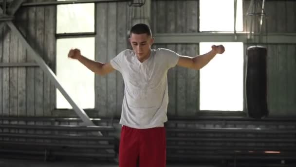 Der junge kaukasische Boxer hüpft und streckt die Arme aus, wärmt sich in der Turnhalle mit herumliegenden Boxsäcken auf. Sport, Boxkonzept — Stockvideo