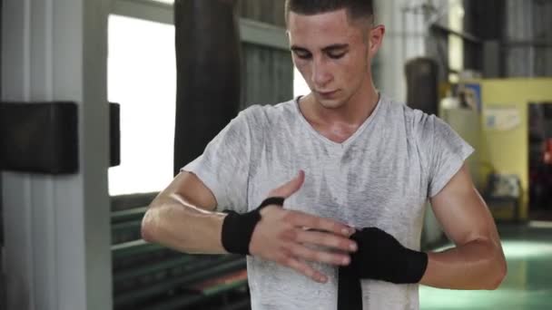 Young, male boxer in boxeng gym preparing for training or competition. Wraps up black bandage on his right fist. Wearing white T shirt and red boxing shorts. Indoors — Stock Video