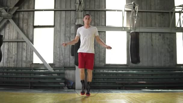Imágenes de cámara lenta de un joven boxeador con camisetas rojas y camiseta haciendo ejercicio con cuerda de salto, haciendo movimientos de pies cruzados. En interiores — Vídeo de stock