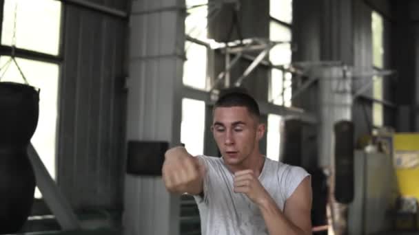 Retrato de un boxeador masculino en camiseta blanca realiza patadas con un oponente invisible, evade, imitando el boxeo. Calentamiento en el gimnasio de boxeo — Vídeos de Stock