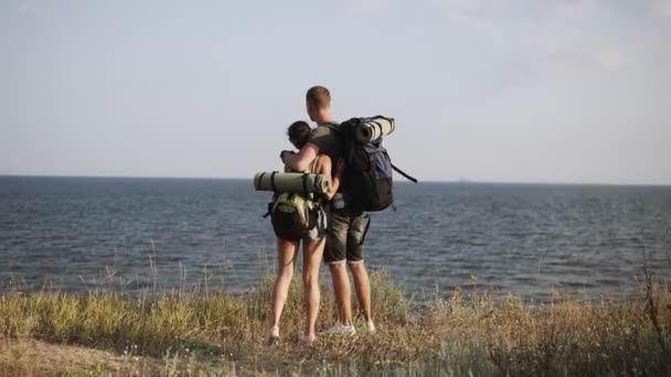 Νεαρό ζευγάρι στέκεται σε αμηχανία στο λόφο με θέα στη θάλασσα. Σακίδιο, πεζοπόρους, ταξιδιώτες. Πίσω προβολή — Αρχείο Βίντεο