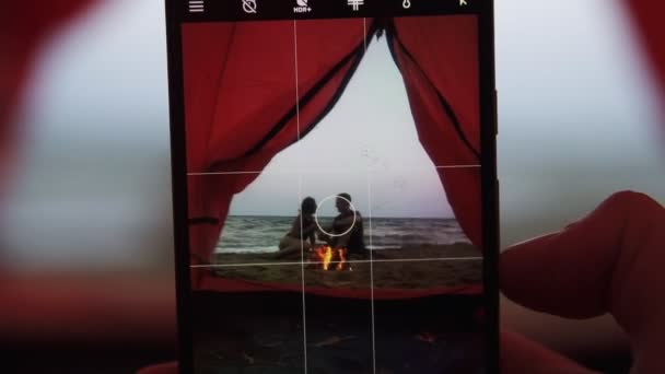 海滩上红色帐篷的录像男性手拍一张年轻浪漫情侣的照片, 坐在火炉旁佛山智能手机。模糊运动效果 — 图库视频影像