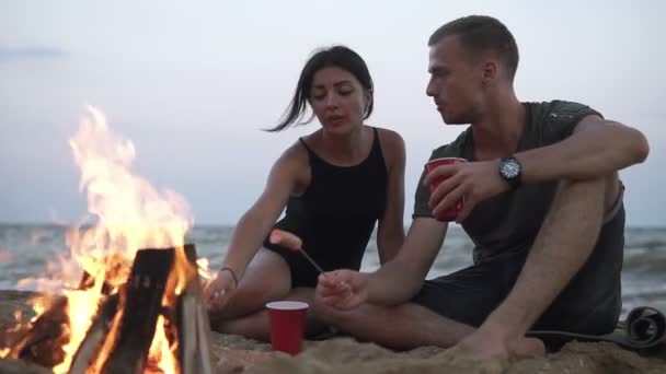 可爱的年轻夫妇花时间在海滩上 穿着黑色泳装的女人 穿着便装的男人用木棍烤香肠 浪漫的时间在一起 谈论篝火附近 — 图库视频影像