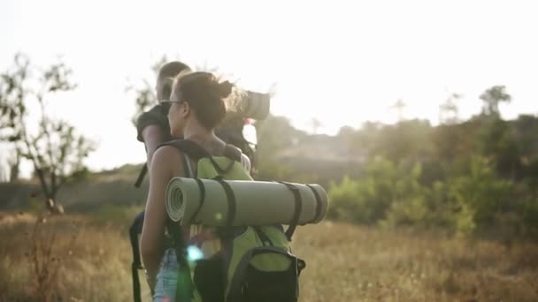 Two travelers - man and woman with huge backpacks are hiking. Walking by grass hills. Sun shines on the background — Stock Video