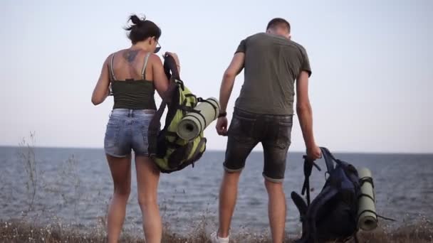 Pareja de senderismo joven descansando en una colina. Mujer joven y hombre excursionista se quitan las mochilas sentadas en el suelo disfrutando de la vista al mar. Pareja joven trekking, relajarse después de la caminata — Vídeos de Stock