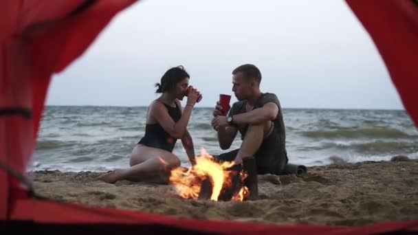 junges Paar - Mann und Frau verbringen Zeit zusammen am Strand in der Nähe des Feuers und trinken aus den roten Plastikbechern. Filmmaterial aus dem roten Zelt. Meer