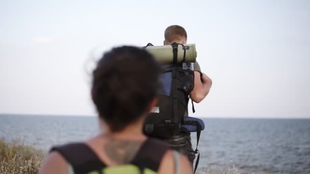 Adventure, travel, tourism, hike and people concept. Front view of a young couple walking with backpacks by hill near the sea. Girl is wearing a sunglasses — Stock Video