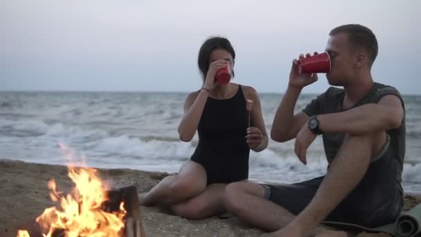 Bella coppia caucasica godendo del tempo sulla spiaggia vicino al falò, bevendo dalle coppe rosse. Donna in costume da bagno nero. Crepuscolo serale — Video Stock