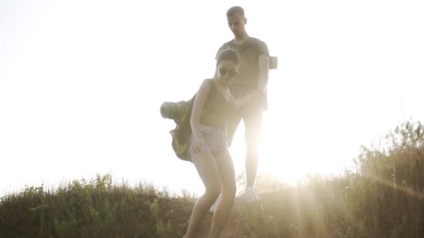 Молодая пара путешественников с рюкзаками, мужчина поддерживает женщину, в то время как она спускается с холма. Солнце светит на заднем плане. Низкий угол съемки — стоковое видео