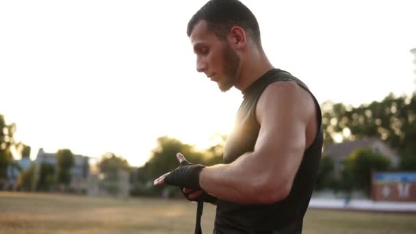 Боковой вид молодого, бородатого боксера, готовящегося к тренировкам на открытом воздухе, заворачивающего черную повязку во время пребывания на стадионе — стоковое видео