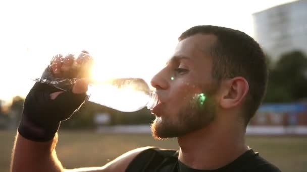 黒包帯で若い男性のボクサーの側ビューは 屋外でトレーニング ペットボトルから水を飲む時に 残りの部分を持ちます 背景に太陽が輝く — ストック動画