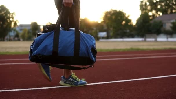 Young muscular man in black shirts walking with blue sport bag in hand by running track of stadium. Ready for training, open bag and take out the boxing gloves. Slow motion — Stock Video