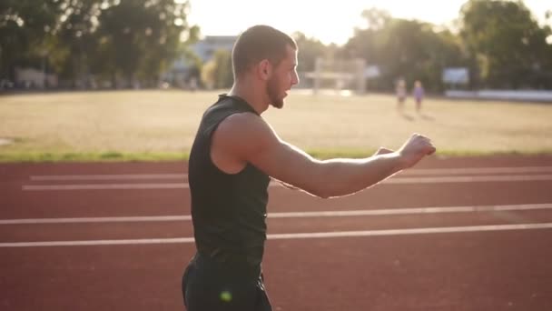 Outdoor shot of young athlete male stretching his hand on a track in stadium. Man wearing black T shirt and shirts. Sun shines on the background — Stock Video